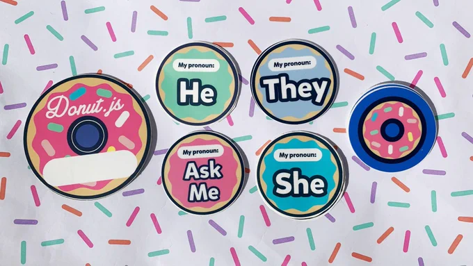 Donut-shaped name tag stickers, Donut.js logo stickers, and several pronoun stickers (Ask Me, He, She, They) arranged on paper patterned with sprinkles.