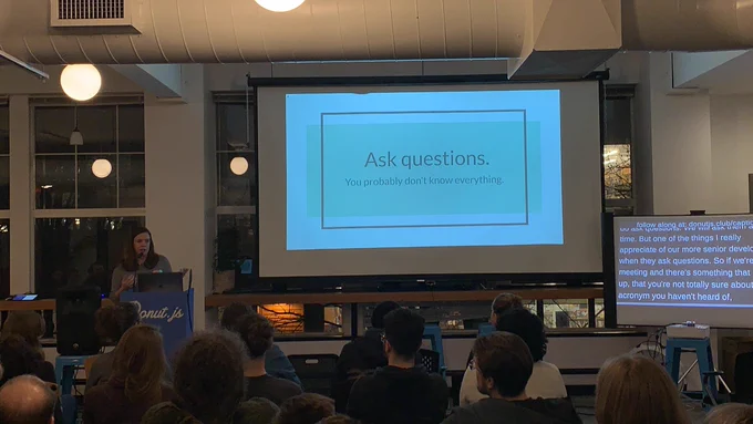 A person giving a talk at a podium in front to a room full of people in chairs. Behind the speaker is a projector screen: “Ask questions. You probably don’t know everything”