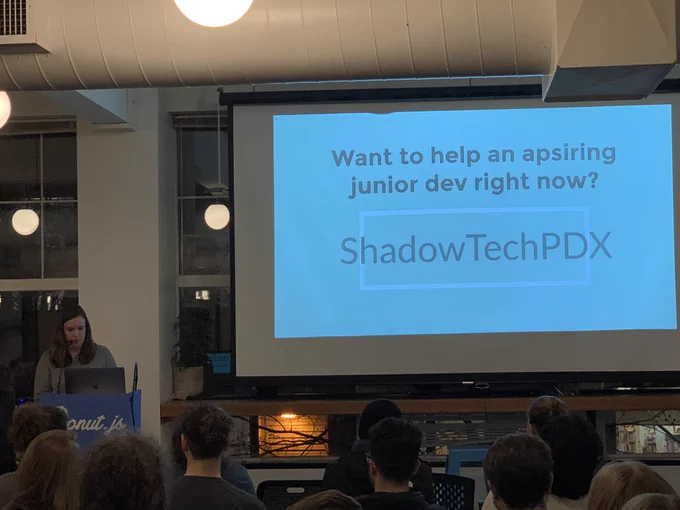 A person giving a talk at a podium in front to a room full of people in chairs. Behind the speaker is a projector screen: “want to help an aspiring junior dev right now? ShadowTextPDX”