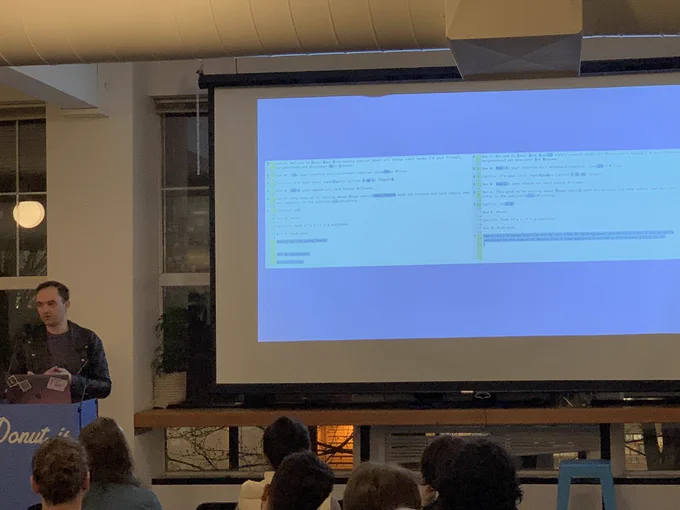 A person giving a talk at a podium in front to a room full of people in chairs. Behind the speaker is a projector screen with a side-by-side diff of an automated transcription fixed up by a person.