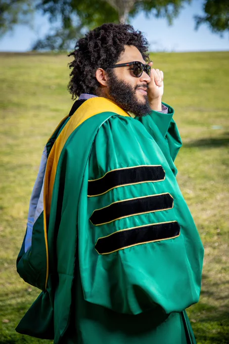 Photo standing in MSU PhD Regalia in grass field showing college hood colors.