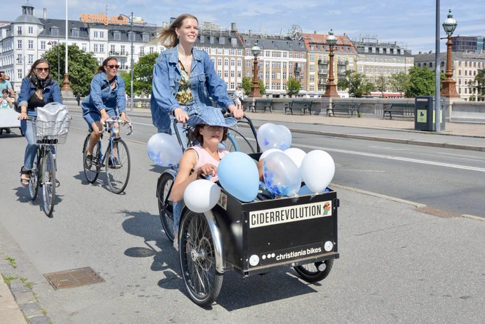 A woman in casual summer attire rides a three-wheeled freight bicycle containing a young woman on the streets of Copenhagen.