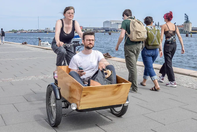 A woman in casual summer attire rides a three-wheeled freight bicycle containing a young man on the streets of Copenhagen.