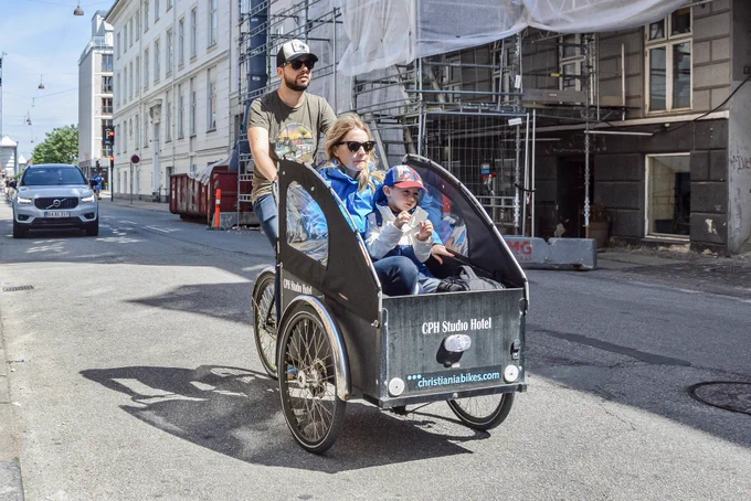 A man in casual summer attire rides a three-wheeled freight bicycle containing a woman and a small child on the streets of Copenhagen.