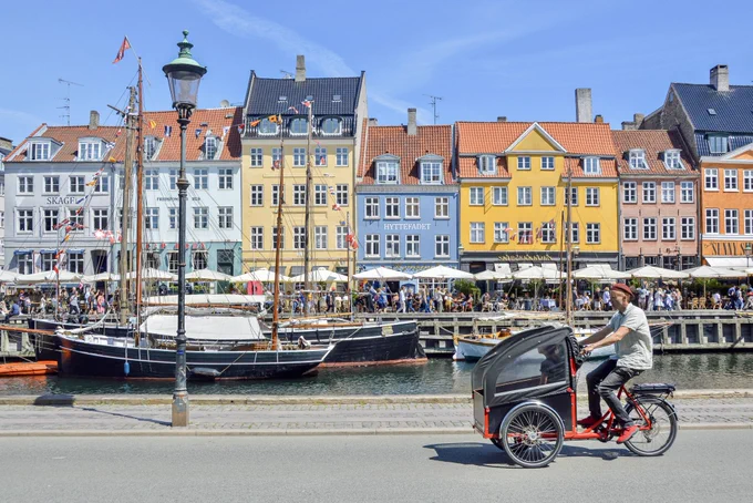 A man in casual summer attire rides a three-wheeled freight bicycle containing a small child on the streets of Copenhagen.