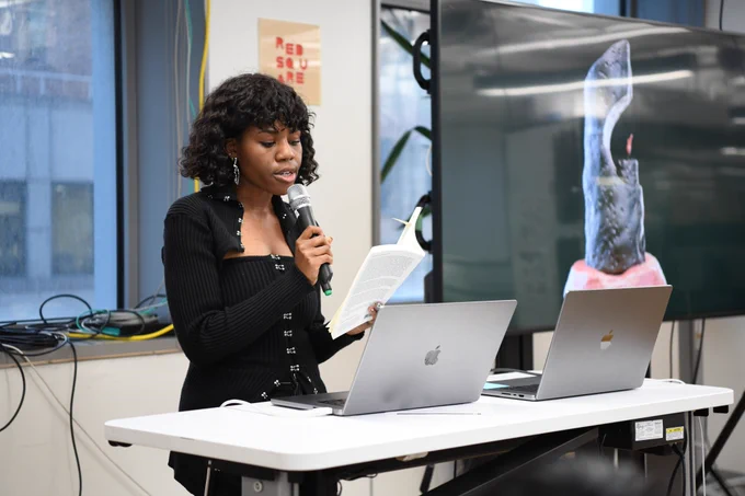An image of Zainab Aliyu holding a mic next to a big t.v. screen that has an image of her work. She is holding a book and reading and excerpt from it
