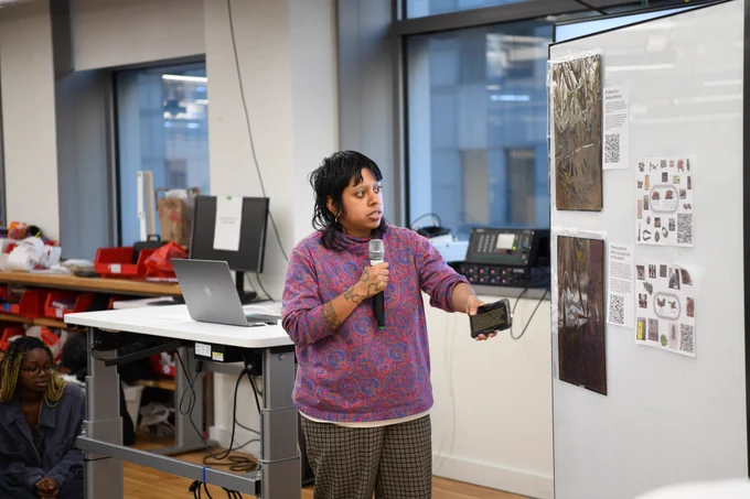 An image of Aarati Akkapedi holding a mic next to a big whiteboard that has a collection of printed work by them. 
