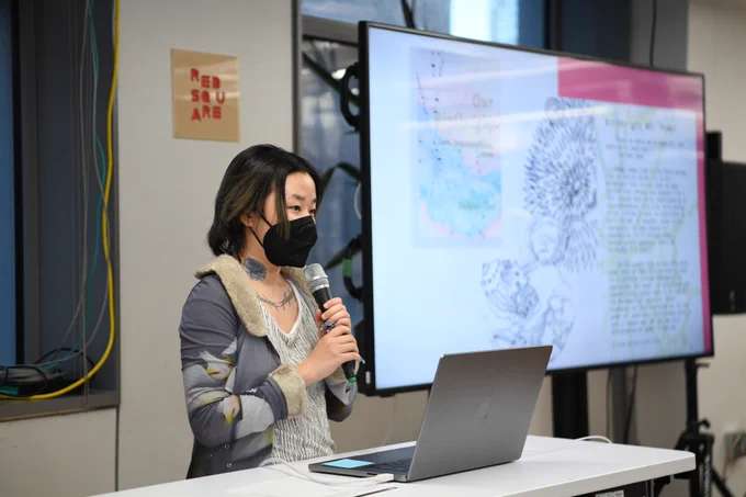 An image of Suhyun Choi holding a mic next to a big t.v. screen that has an image of their work. 

