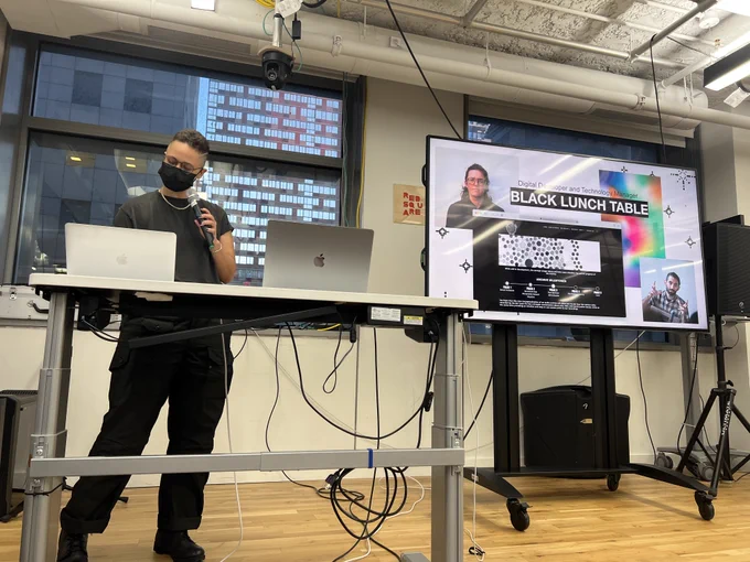 An image of Shawné Michaelain Holloway holding a mic next to a big t.v. screen that has an image of their work called, ‘Black Lunch Table’. 
