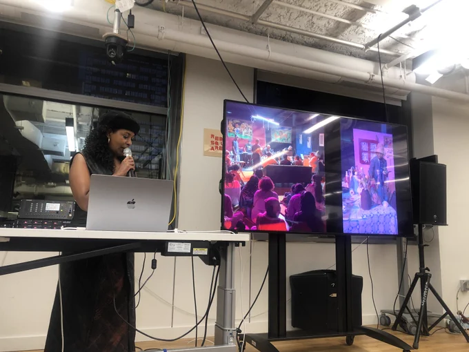 An image of Tsige Tafesse holding a mic next to a big t.v. screen that has an image of their work from MoCADA. 
