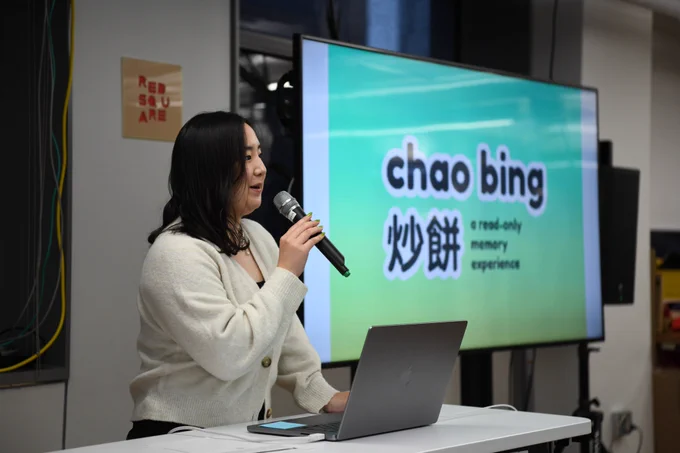 An image of Jackie Liu holding a mic next to a big t.v. screen that has the title of her presentation and work, “Chao bing” written on it in English and Chinese.
