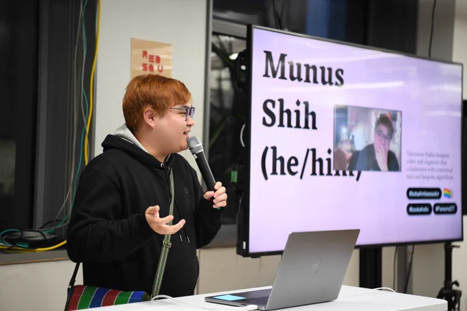 An image of Munus Shih holding a mic next to a big t.v. screen that has an image of himself with his name, pronouns, and other facts about him
