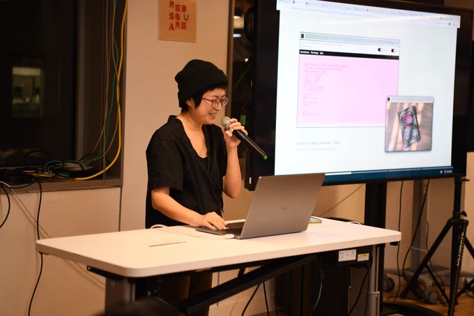 An image of Xin Xin holding a mic next to a big t.v. screen that has an image and text of their work
