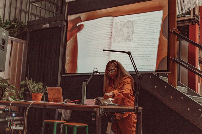 An image of Kemi Ukadike reading the Processing Community Catalog at Hex House. The image of the book is projected onto the projector screen behind Kemi Ukadike as live footage. Their hand is touching the book and keeping it open to its page, while holding a mic on the other hand.
