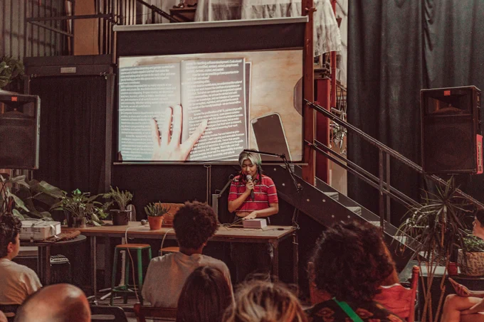 An image of Viola He reading the Processing Community Catalog at Hex House. The image of the book is projected onto the projector screen behind Viola He as live footage. Their hand is touching the book and keeping it open to its page, while holding a mic on the other hand. In the front of the image are a couple of audience members.
