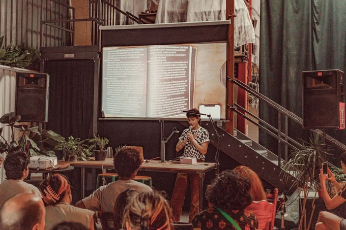 An image of Cassie Tarakajian reading the Processing Community Catalog at Hex House. The image of the book is projected onto the projector screen behind Cassie Tarakajian as live footage. Their hand is on their chest while holding a mic on the other hand. In the front of the image are a couple of audience members.

