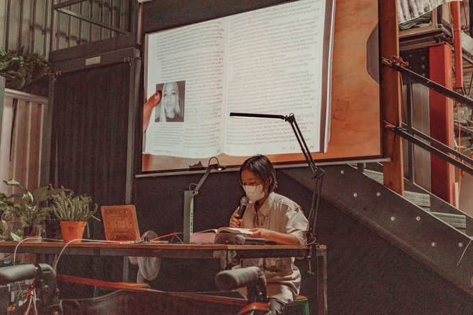 An image of Suhyun Choi reading the Processing Community Catalog at Hex House. The image of the book is projected onto the projector screen behind Suhyun Choi as live footage. Their hand is touching the book and keeping it open to its page, while holding a mic on the other hand. 
