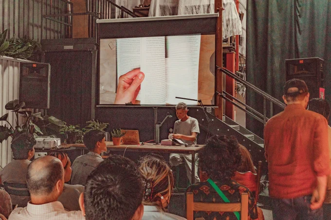 An image of Casey Reas reading the Processing Community Catalog at Hex House. The image of the book is projected onto the projector screen behind Casey Reas as live footage. Their hand is touching the book and keeping it open to its page, while holding a mic on the other hand. In the front of the image are a couple of audience members.
