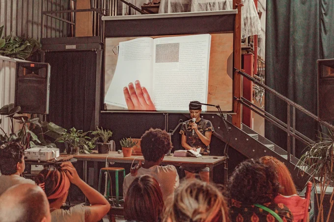 An image of Lauren Lee McCarthy reading the Processing Community Catalog at Hex House. The image of the book is projected onto the projector screen behind Lauren Lee McCarthy as live footage. Their hand is touching the book and keeping it open to its page, while holding a mic on the other hand. In the front of the image are a couple of audience members.
