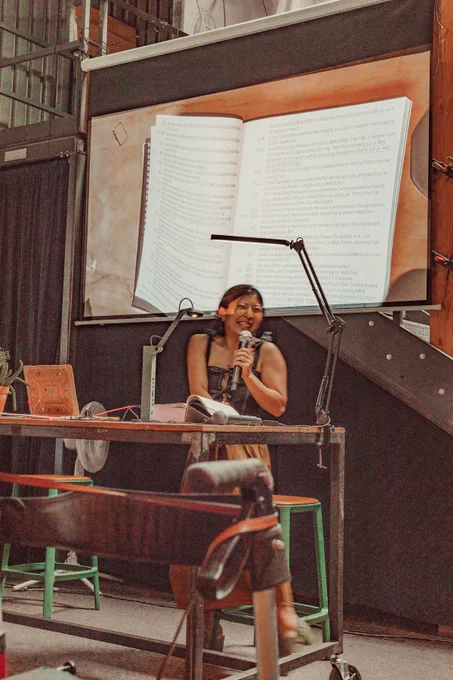 An image of Rachel Lim reading the Processing Community Catalog at Hex House. The image of the book is projected onto the projector screen behind Rachel Lim as live footage. Rachel is smiling mid-laughter and is about to leave the reading area.
