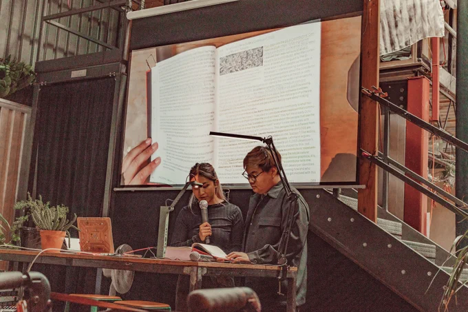 An image of Munus and Nikki reading the Processing Community Catalog at Hex House. The image of the book is projected onto the projector screen behind Munus and Nikki as live footage. Their hand is touching the book and keeping it open to its page, while holding a mic on the other hand.
