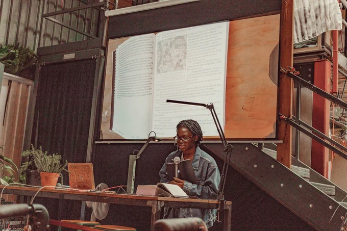 An image of Olivia McKayla Ross reading the Processing Community Catalog at Hex House. The image of the book is projected onto the projector screen behind Olivia McKayla Ross as live footage.
