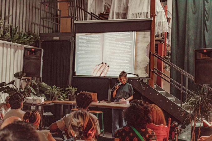 An image of Tega Brain reading the Processing Community Catalog at Hex House. The image of the book is projected onto the projector screen behind Tega Brain as live footage. Their hand is touching the book and keeping it open to its page, while holding a mic on the other hand. In the front of the image are a couple of audience members.
