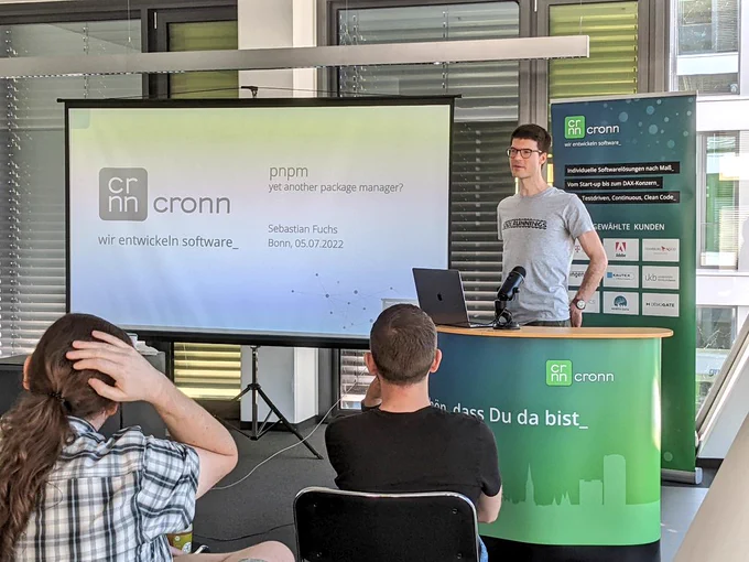 Sebastian on stage in our auditorium. Behind  him on screen there's the company logo of cronn and the title of the talk "pnpm - yet another package manager"