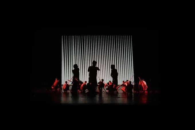 Scena di teatro con personaggi seduti per terra con tronchi. Sul fondale digitale tre grandi sagome che discutono. 