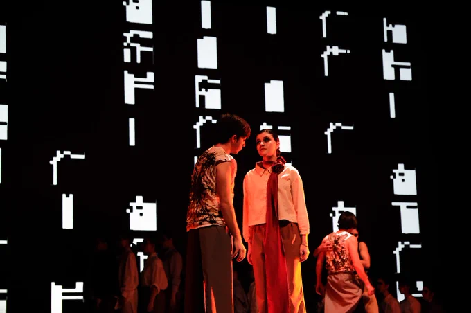 Scena di teatro con ragazzi che si guardano, fondale digitale con caratteri tipografici astratti. 