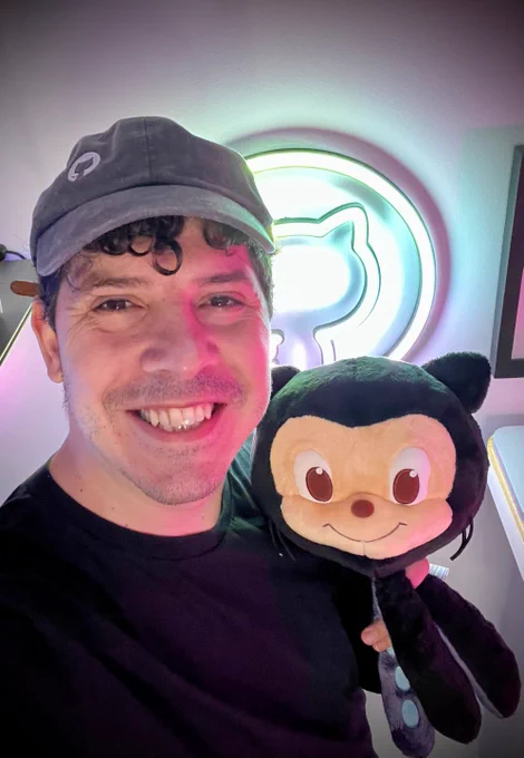Aparezco sonriendo a cámara con mi peluche y mi gorra de GitHub. Al fondo hay unas luces de neón con el logo de GitHub.