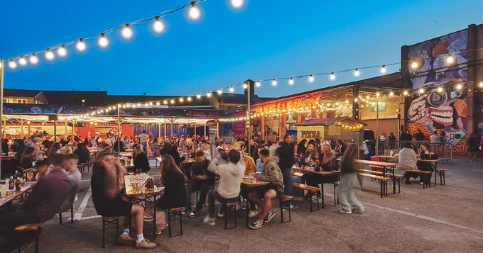 Picture of Luna Springs. Open air space with long tables and benches with people sat down drinking and talking. 