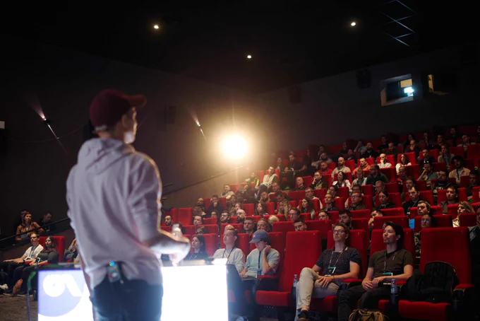 Me, wearing a pink Deno hoodie, a speaker badge, and a red cap. I am standing in front of an audience sitting in a movie theatre, giving a talk.
