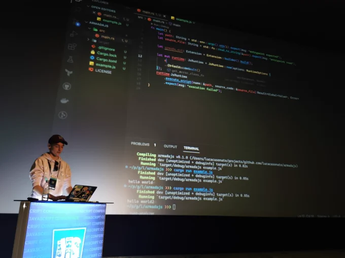 Me, wearing a pink Deno hoodie, a speaker badge, and a red cap. I am standing in front of a screen giving a talk.