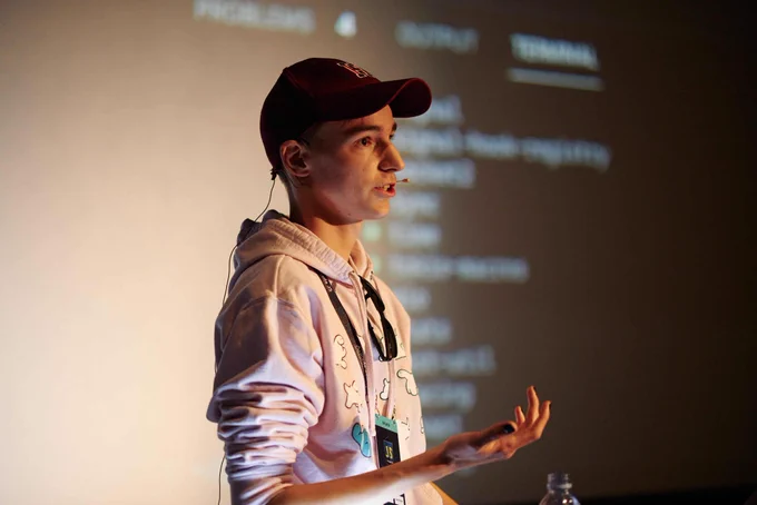Me, wearing a pink Deno hoodie, a speaker badge, and a red cap. I am standing in front of a screen giving a talk.