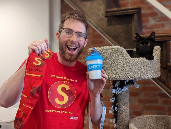 Me smiling, wearing a Stryker Mutation Testing shirt, holding a Stryker shirt and stickers, next to a black cat on a cat tower with Halloween decoration garland