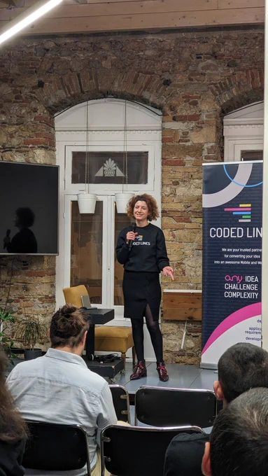 A woman wearing black on the stage of GreeceJS meetup presenting our sponsor for the 48th meetup Coded Lines, an end-to-end mobile and  web app development and digital marketing agency.