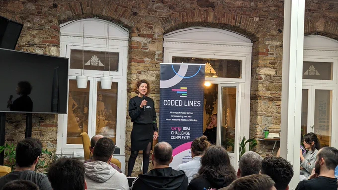 A woman wearing black on the stage of GreeceJS meetup presenting our sponsor for the 48th meetup Coded Lines, an end-to-end mobile and  web app development and digital marketing agency.