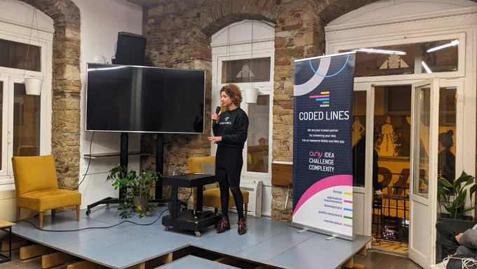 A woman wearing black on the stage of GreeceJS meetup presenting our sponsor for the 48th meetup Coded Lines, an end-to-end mobile and  web app development and digital marketing agency.