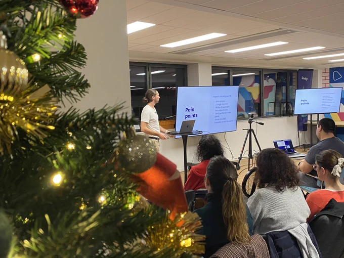 Romain Koenig parle devant le public de LyonJS de Trunk Based Development chez Indy. Un sapin de Noël est au premier plan.