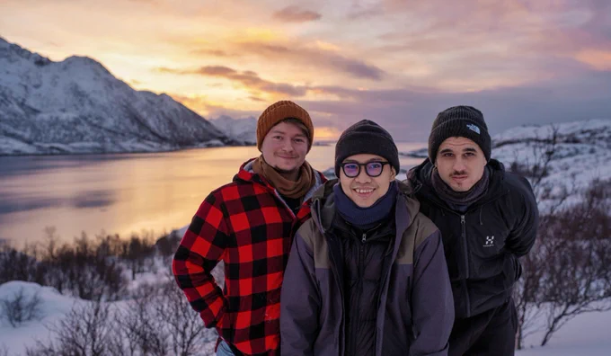 Scenic sunrise in Lofoten Islands, Norway, with a snowy mountain on the left of the image, and the ocean behind us, with some snowy landscape as well. In the middle, from left to right, Balázs, Thang and Lluis facing the camera, with a slight smile.