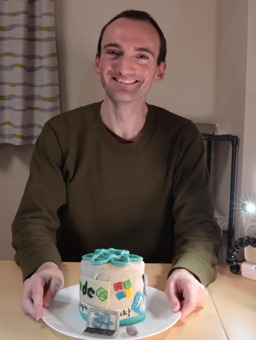 A pleased Jamie holding his surprise birthday cake, which is adorned with marzipan logos for Node.js, Microsoft, Expo, Callstack, NativeScript, and React Native.