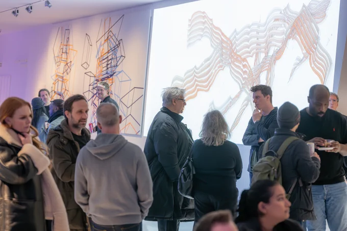 A group of people is gathered at an art exhibition, engaging in conversations and observing digital and geometric artwork displayed on the walls. The exhibition space is illuminated with soft lighting. A large digital screen in the background showcases abstract, flowing patterns in white and orange, while another artwork features geometric line structures in black, orange, and blue. Attendees are dressed in warm clothing, with some holding drinks, while others appear deep in discussion or contemplation of the art.