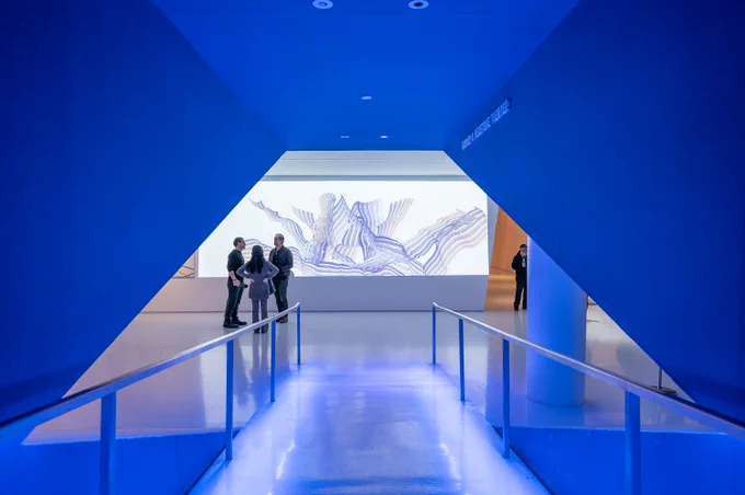 A modern art exhibition space with a blue-lit entrance leading to a well-lit gallery. In the background, a large digital artwork with abstract, flowing patterns in white and blue is displayed on a screen. Three people are standing in front of the artwork, engaged in conversation, while another person stands near the wall observing the exhibit. The scene is framed by geometric blue walls, creating a futuristic ambiance.