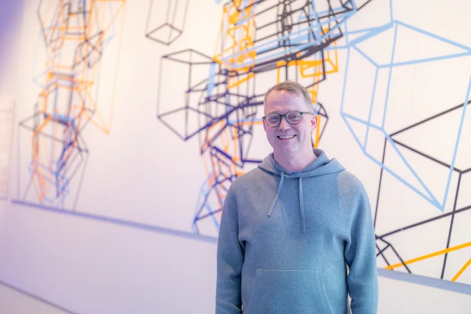 Marius Watz, wearing glasses and a gray hoodie, stands in front of a geometric artwork displayed on a white gallery wall. The artwork consists of interconnected black, blue, and orange lines forming abstract cubic structures. The exhibition space is well-lit with a modern, minimalist aesthetic. 