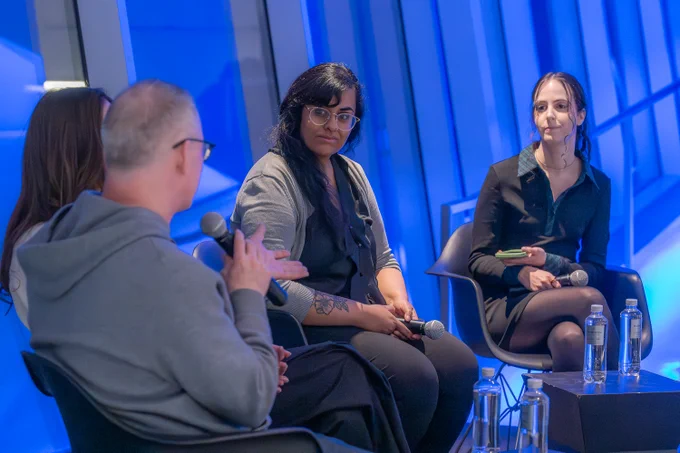  A panel discussion is taking place in a modern venue with blue lighting. Four individuals are seated in black chairs, engaged in conversation. Marius Watz is in the foreground, wearing glasses and a gray hoodie. Roopa Vasudevan sits in the middle, wearing glasses, a black top, and a gray cardigan. On the right, Regina Harsanyi holds a notepad and microphone while looking towards the speaker. Water bottles are placed on a table in front of them. The background features large windows with reflective blue panels.