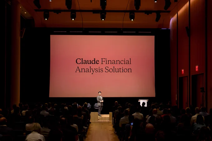 A presenter on stage in front of a large audience in an auditorium. Behind them is a large projection screen displaying "Claude Financial Analysis Solution."