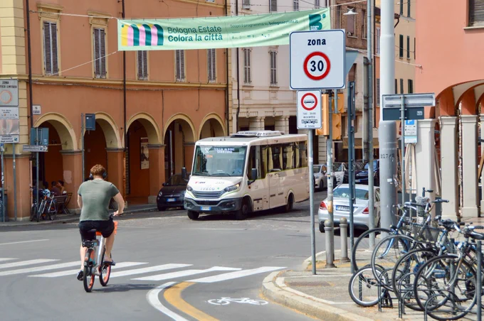 People of all ages and abilities walk, cycle, drive, and ride public transportation along the low-traffic streets of Bologna, which are lined with buildings, trees, bike infrastructure, and prominent 30 km/h speed limit signage and road markings.