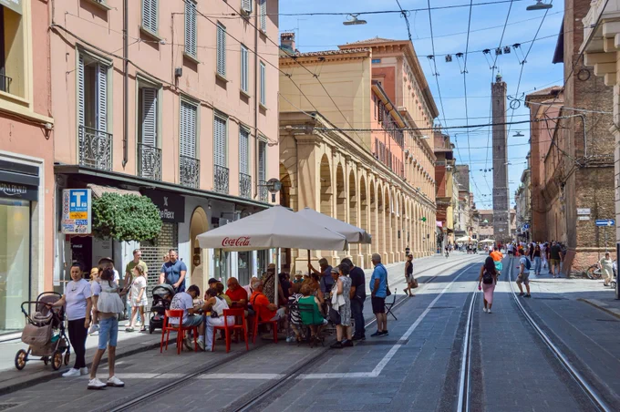 People of all ages and abilities walk, cycle, drive, and ride public transportation along the low-traffic streets of Bologna, which are lined with buildings, trees, bike infrastructure, and prominent 30 km/h speed limit signage and road markings.